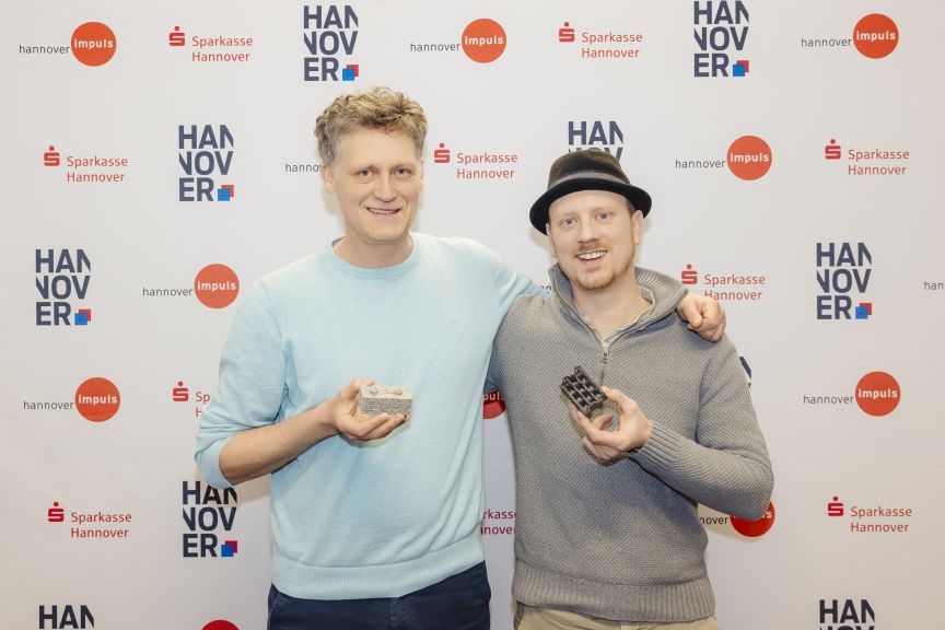 Zwei Männer stehen vor einer Sponsorenwand mit Hannover-Impuls und Sparkasse-Logos und halten kleine Objekte in den Händen.