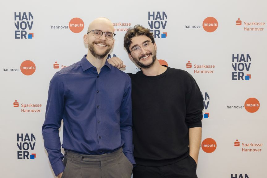 Zwei junge Männer mit Brillen lächeln vor einer Sponsorenwand mit Logos von Hannover Impuls und Sparkasse Hannover.
