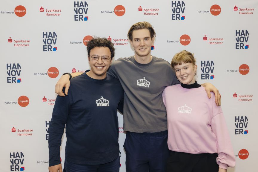 Drei Personen posieren lächelnd vor einer Hannover-Impuls-Sparkasse-Wand. Alle tragen Sweatshirts mit „Renew Merch"-Logo.