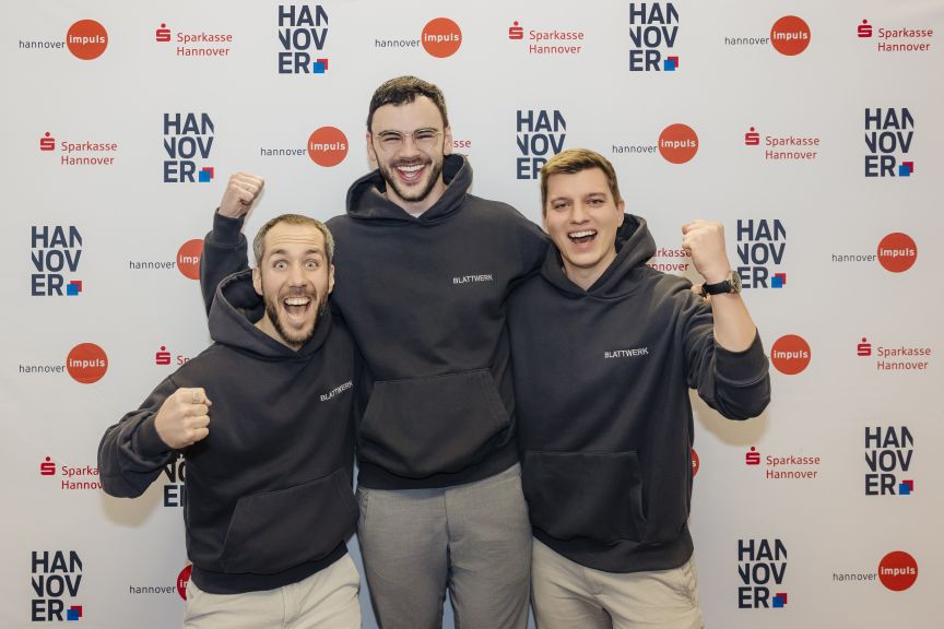 Drei junge Männer in schwarzen Hoodies posieren jubelnd vor einer Hannover-Impuls/Sparkasse-Hannover-Werbewand.
