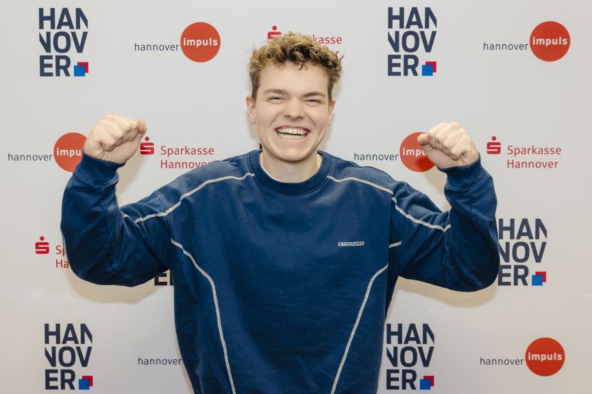 Junger Mann in blauem Sweatshirt ballt jubelnd die Fäuste vor Hannover-Impuls- und Sparkasse-Hannover-Banner.