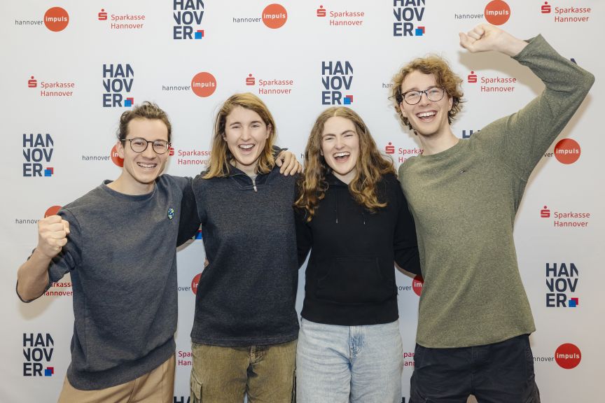 Vier junge Menschen stehen lachend vor einer Sponsorenwand mit Logos von Sparkasse Hannover und Hannover Impuls.