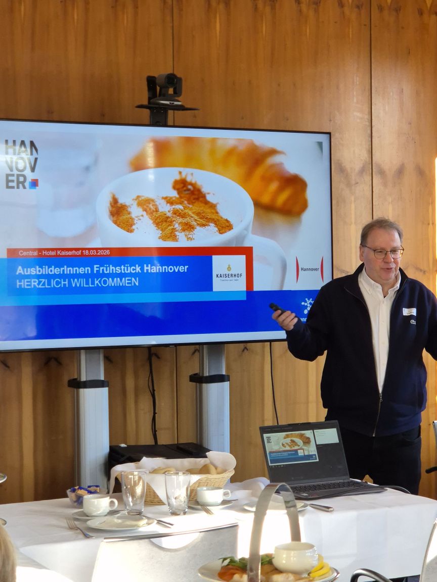 Mann steht vor Präsentationsfolie „AusbilderInnen Frühstück Hannover – Herzlich Willkommen" im Hotel Kaiserhof.