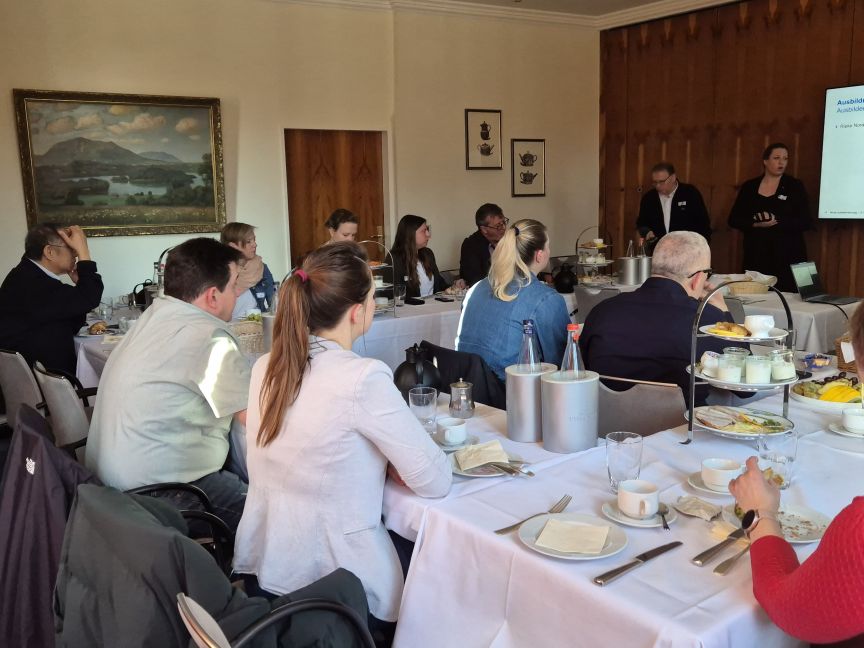 Gruppe von Personen sitzt an gedeckten Tischen in einem Konferenzraum; zwei Personen stehen vor einer Präsentation.