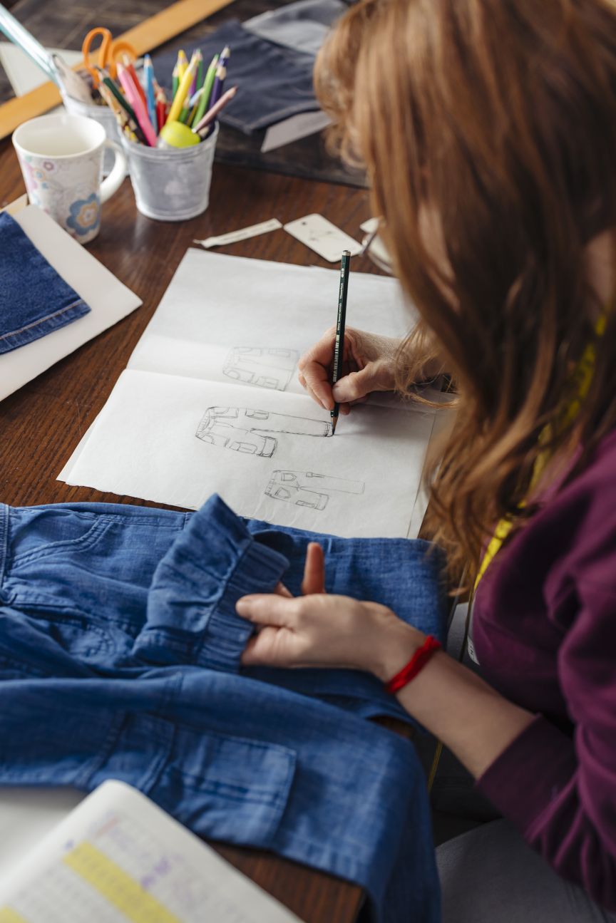 Frau mit roten Haaren zeichnet Skizzen auf Papier, hält Jeanshose, Tisch mit Stiften und Scheren.