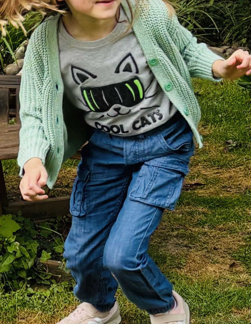 Kind in mintgrüner Strickjacke, grauem „Cool Cats"-Shirt und blauen Cargojeans läuft auf Rasen.