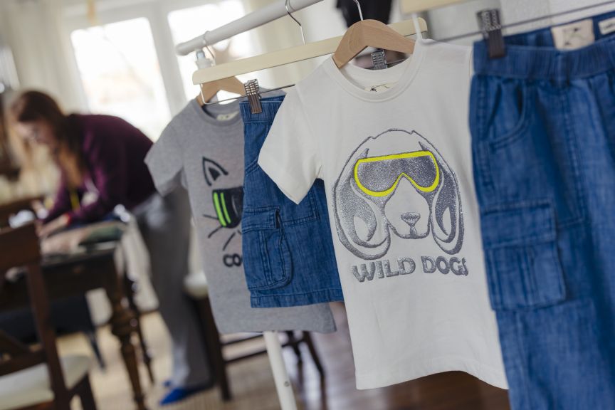 Kinderkleidung auf Bügeln: weißes T-Shirt „Wild Dogs", graues T-Shirt, Jeansrock, Jeanshose. Im Hintergrund eine Person.