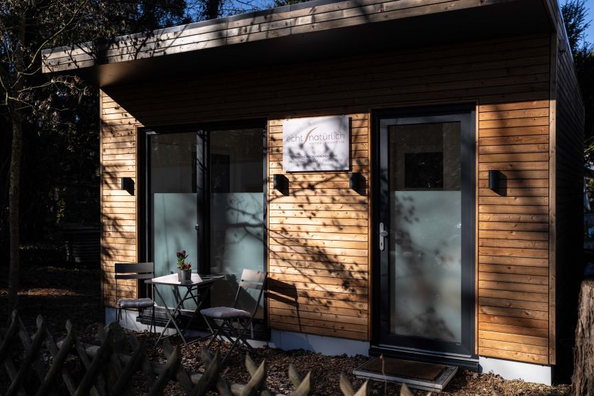Modernes Holzgebäude mit Glasfront, kleinem Bistrotisch und zwei Stühlen davor, umgeben von Bäumen.