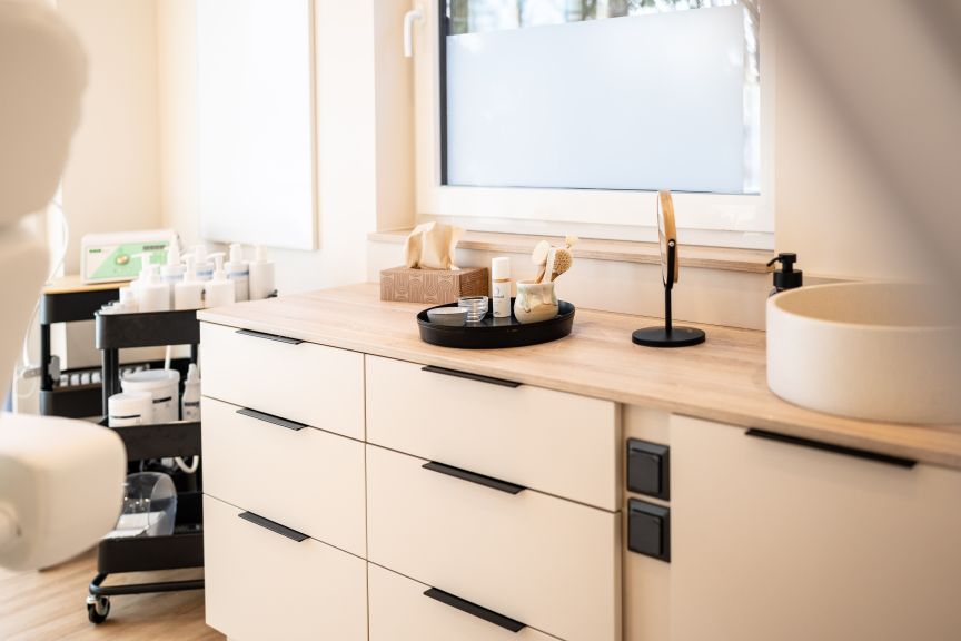 Helle Kommode mit schwarzen Griffen, Tray mit Kosmetikprodukten, Waschbecken und Rollwagen mit Flaschen im Hintergrund.