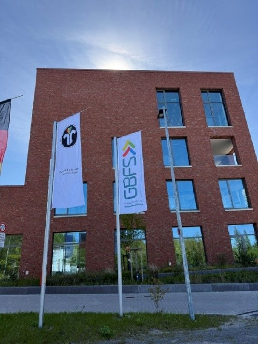Rotes Backsteingebäude mit zwei Fahnen: Logo mit "r" und "GBFS"-Schriftzug. Blauer Himmel mit Sonnenschein.