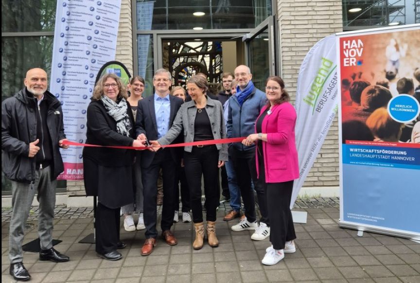 Gruppe von Personen bei Eröffnungsfeier mit rotem Band vor Gebäude in Hannover, Bannern und Fahnen im Hintergrund.