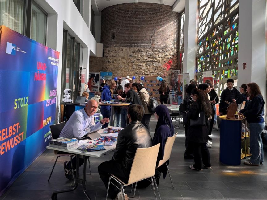 Messestand mit Besucher:innen in einer Halle; buntes Banner mit deutschen Wörtern wie „Stolz", „Happy", „Selbstbewusst".