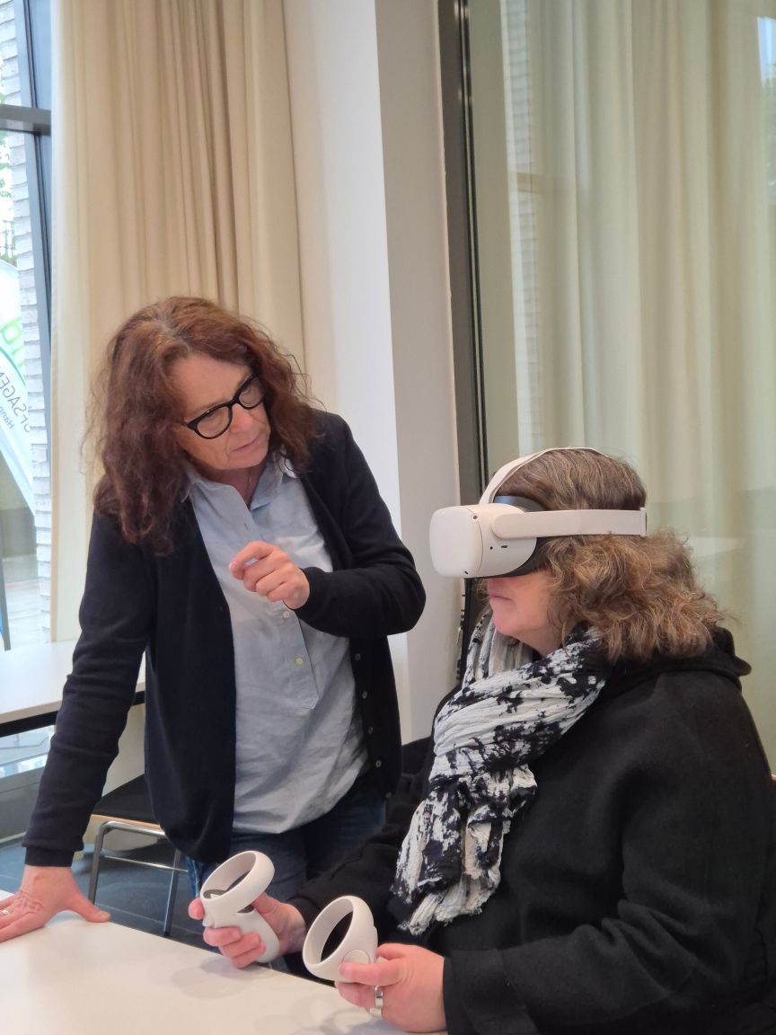 Zwei Frauen, eine stehend mit Brille, eine sitzend mit VR-Headset und Controllern in den Händen.