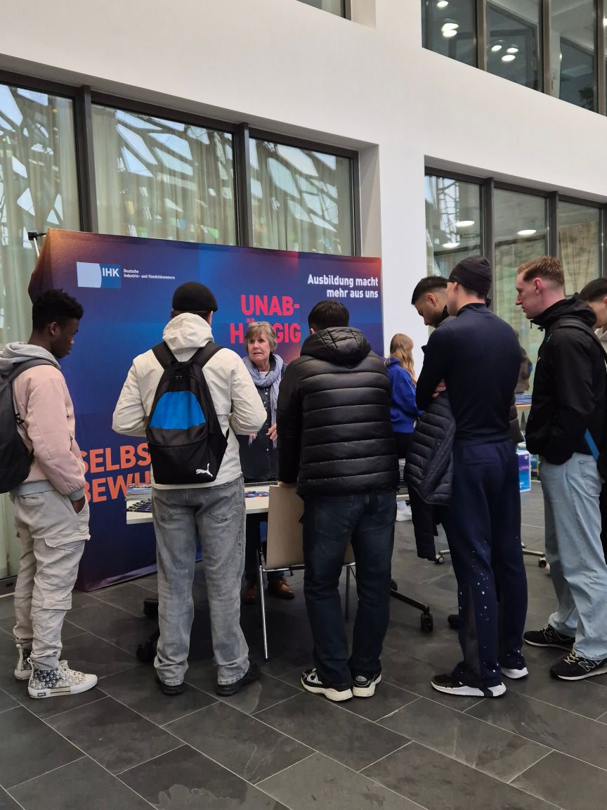 Jugendliche stehen an einem IHK-Informationsstand mit Bannern zu Ausbildung in einer modernen Halle.