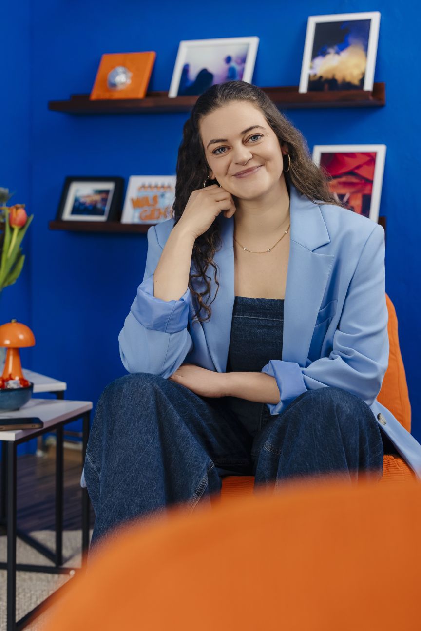 Junge Frau mit lockigem Haar in hellblauem Blazer sitzt lächelnd vor blauer Wand mit Bilderregalen.