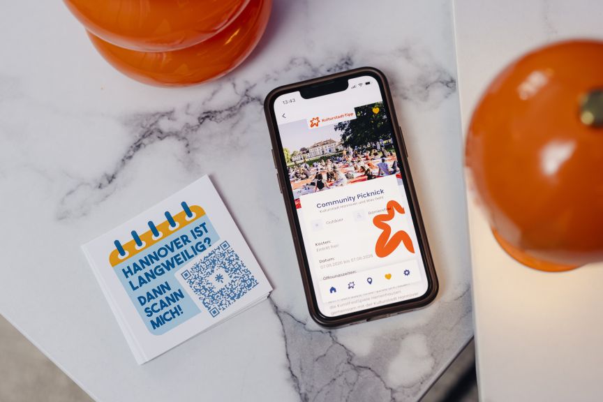 Smartphone zeigt App mit "Community Picknick", daneben Flyer mit QR-Code und Text "Hannover ist langweilig? Dann scann mich!"