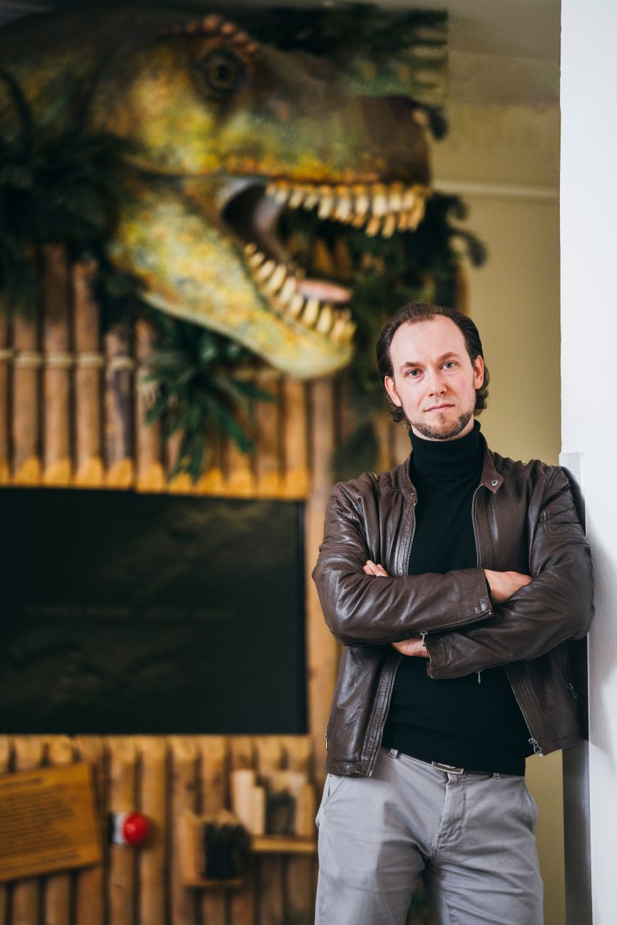 Mann mit Lederjacke und verschränkten Armen steht vor einem großen T-Rex-Modell in einem Museum.