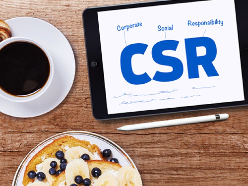 Tablet mit "CSR – Corporate Social Responsibility"-Anzeige, daneben Kaffee, Croissant, Obstteller und Stift auf Holztisch.