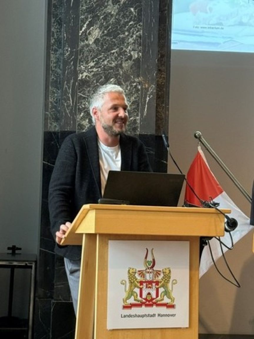 Mann mit grauem Bart am Rednerpult der Landeshauptstadt Hannover mit Laptop, lächelnd, im Hintergrund Präsentation.