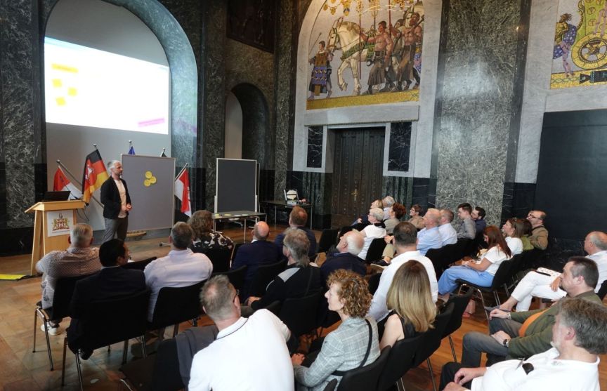 Ein Mann präsentiert vor Publikum in einem Saal mit Wandgemälden und Flaggen, dahinter eine Leinwand mit Folien.