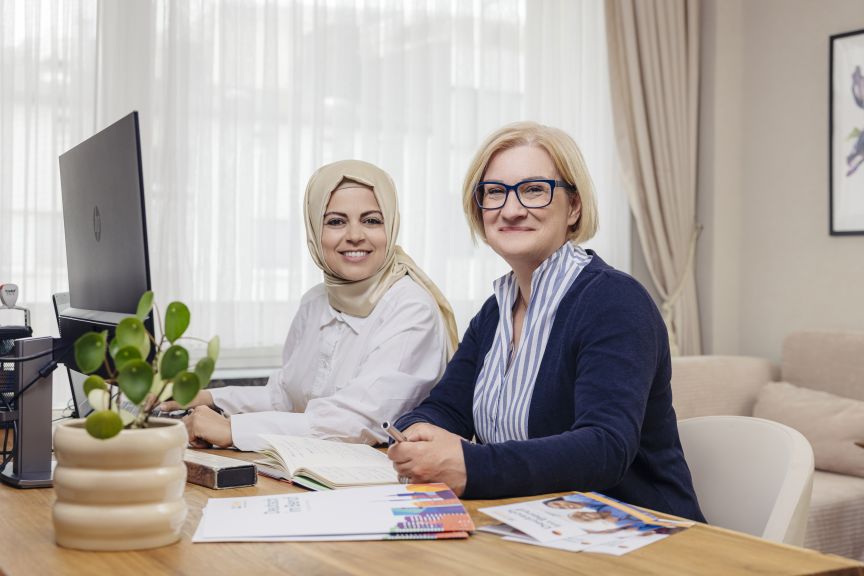 Zwei Frauen sitzen an einem Schreibtisch mit Computer, Dokumenten und Pflanze; eine trägt Hijab, die andere Brille.