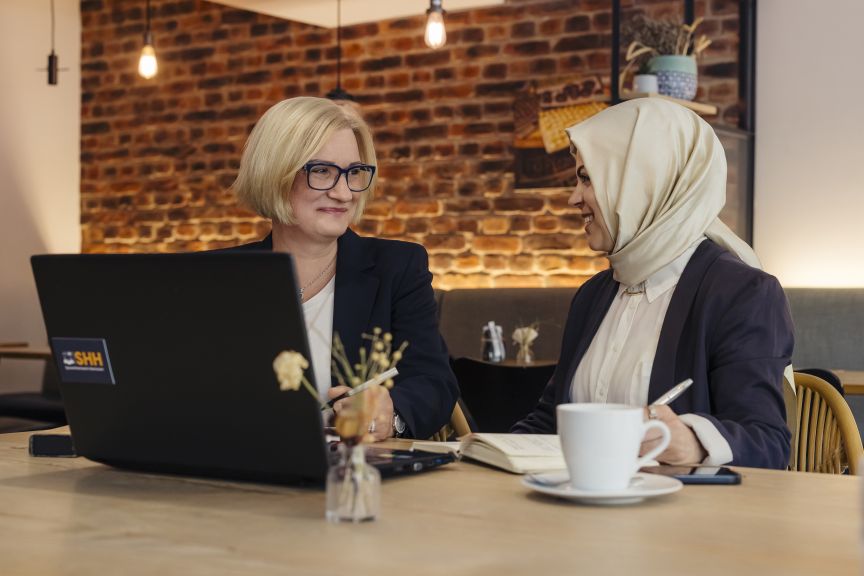 Zwei Frauen sitzen an einem Tisch mit Laptop und Notizbuch. Eine trägt Hijab, die andere eine Brille.