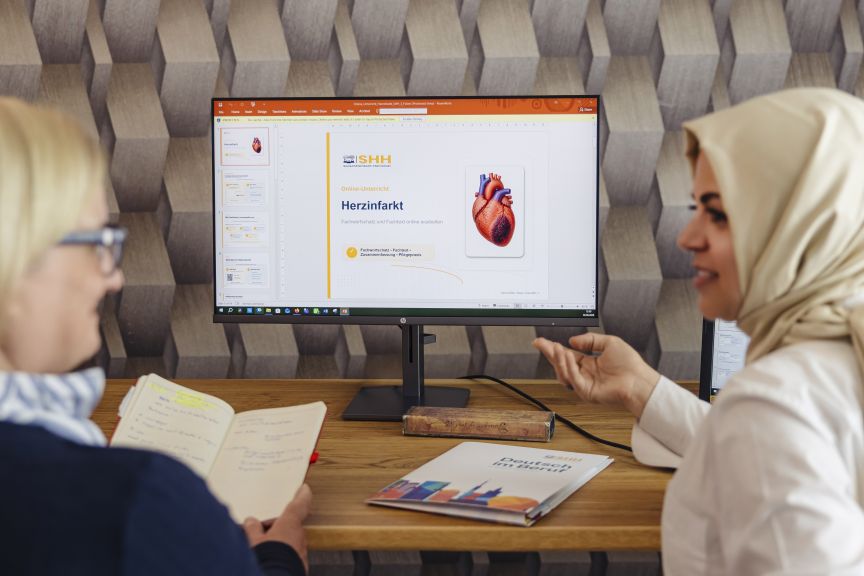 Zwei Frauen vor Monitor mit PowerPoint-Präsentation zum Thema „Herzinfarkt" mit Herzanatomie-Grafik.