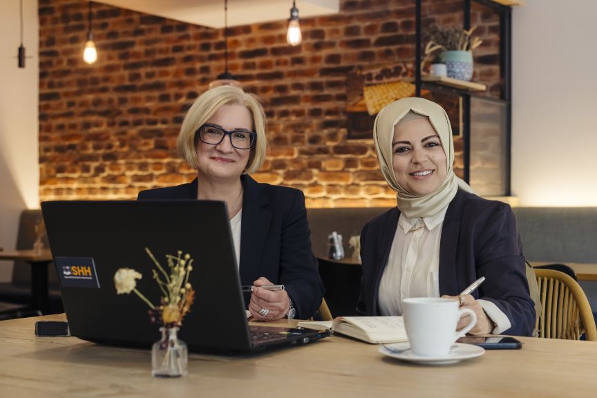 Zwei Frauen sitzen an einem Holztisch mit Laptop, Notizbuch und Kaffeetasse und lächeln in die Kamera.