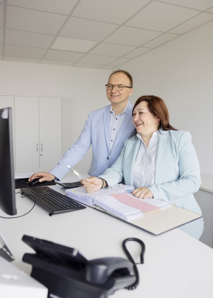 Zwei Personen in hellen Blazern an einem Schreibtisch mit Computer, Tastatur und Aktenordner, lachend.