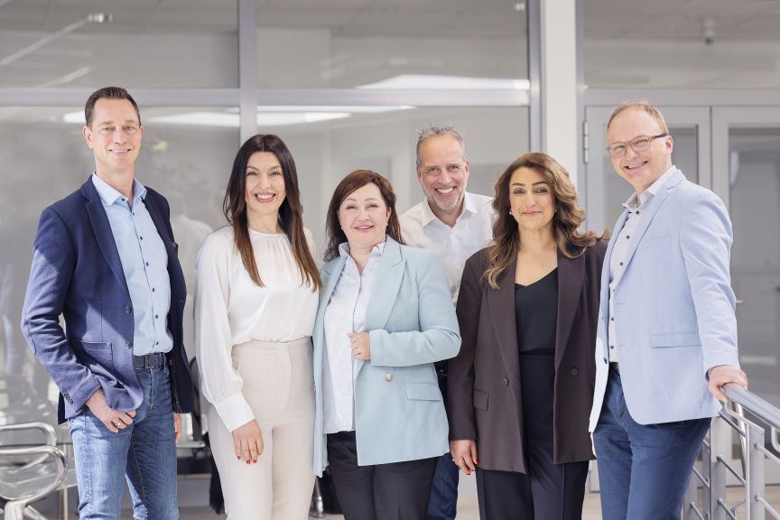 Sechs Personen, drei Männer und drei Frauen, stehen lächelnd nebeneinander in einem modernen Bürogebäude.
