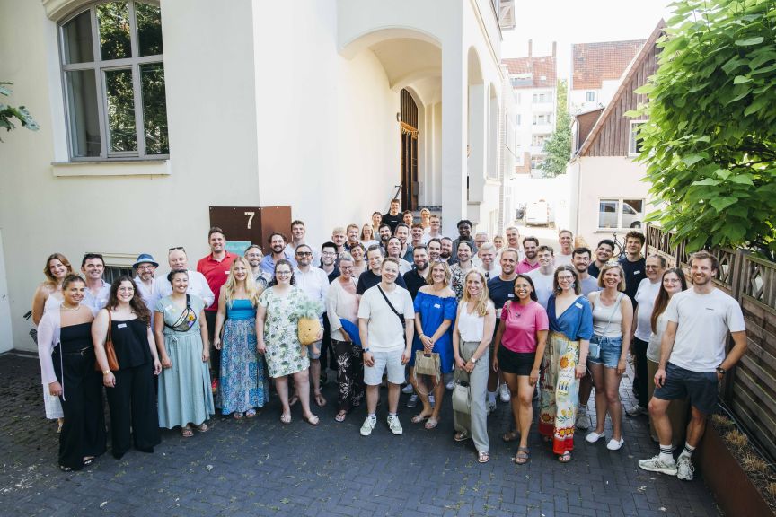 Große Gruppe von etwa 60 Personen steht vor einem hellen Gebäude mit der Hausnummer 7, sommerlich gekleidet.