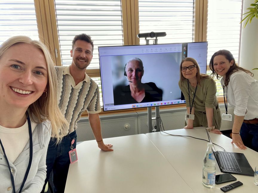 4 Personen und eine eine Person auf einem Bildschirm innerhalb einer Videokonferenz. Alle schauen in die Kamera und lächeln.