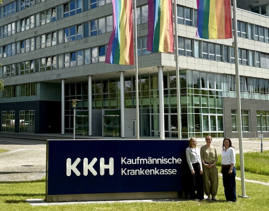 Drei Frauen vor dem KKH Bürogebäude, Regenbogenflaggen wehen im Hintergrund.
