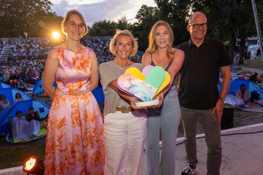 Drei Frauen und ein Mann mit dem Preis auf der Bühne. Im Hintergrund ist Publikum.