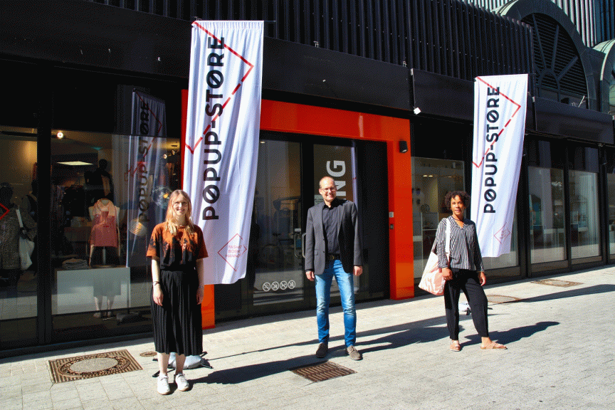 Drei Personen stehen vor einem Popup-Store mit weißen Fahnen und orangefarbenem Eingang.