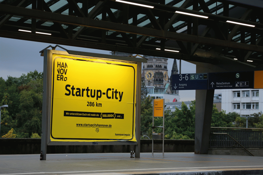 Gelbes Werbeschild „Hannover Startup-City 286 km" auf einem Bahnsteig, im Hintergrund Gebäude und Bahnhofsbeschilderung.