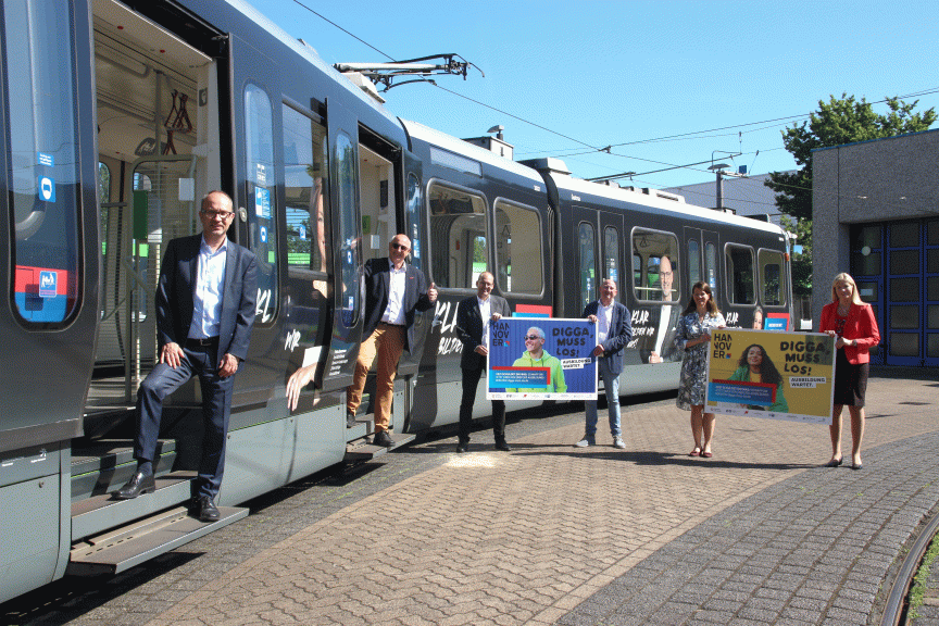 Gruppe von sieben Personen steht vor einer modernen Straßenbahn und hält Werbeplakate mit der Aufschrift „Digga muss los".