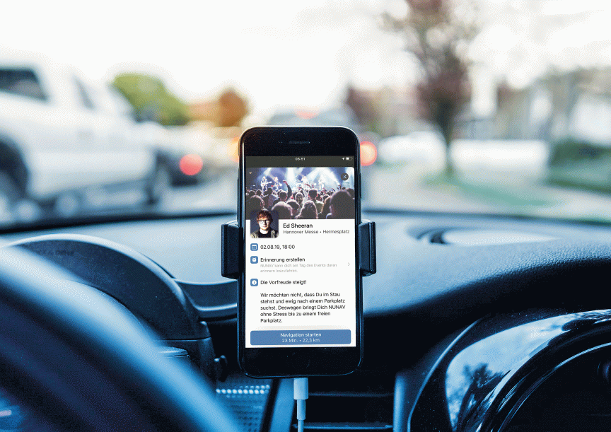 Smartphone in KFZ-Halterung auf Armaturenbrett zeigt Event-App mit Ed-Sheeran-Konzertinfo, Straße im Hintergrund.