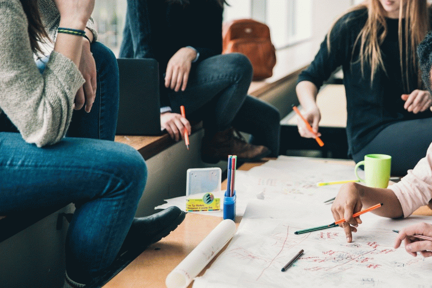 Mehrere Personen sitzen um einen Tisch mit großem Papier, Stiften und einer grünen Tasse und arbeiten gemeinsam.