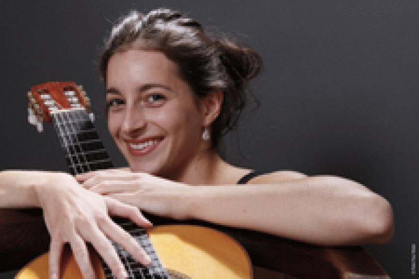 Lächelnde Frau lehnt mit verschränkten Armen auf einer Akustikgitarre vor dunklem Hintergrund.