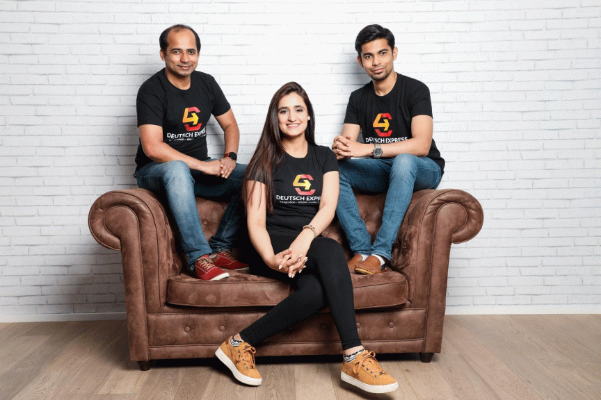 Drei Personen in schwarzen „Deutsch Express"-T-Shirts posieren auf und vor einem braunen Ledersofa vor einer Backsteinwand.
