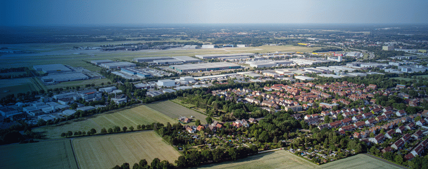 Luftaufnahme einer Stadt mit Wohngebiet, Industriegebäuden, landwirtschaftlichen Feldern und flacher Umgebungslandschaft.