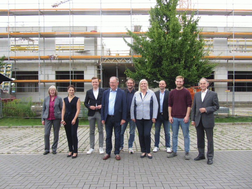 Gruppe von 10 Personen steht vor einem Gebäude im Rohbau mit Baugerüst und einem Baum.