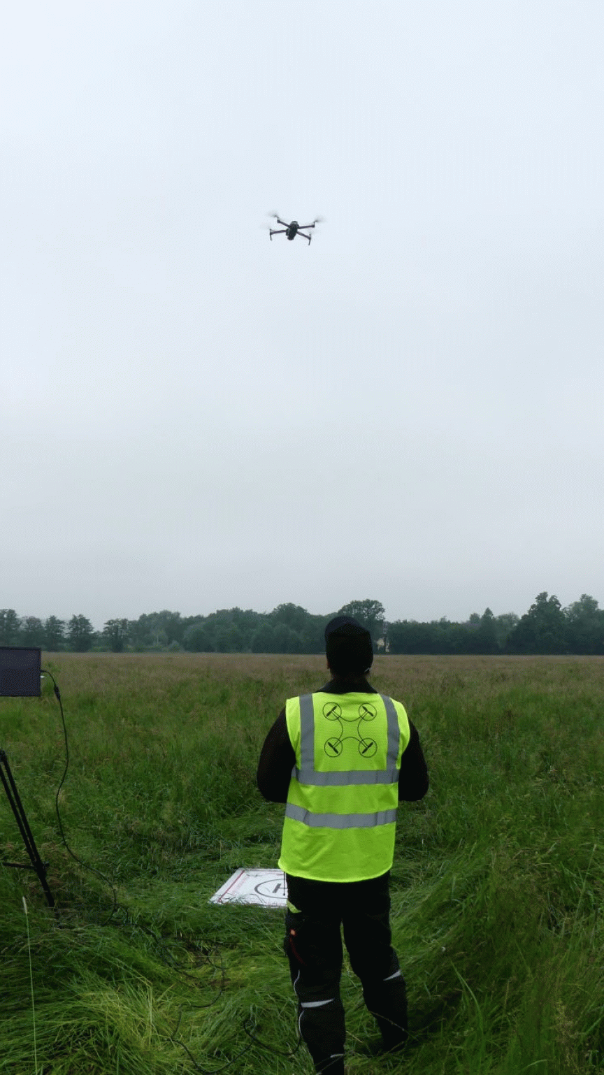 Person in gelbер Warnweste steht auf einer Wiese und steuert eine Drohne, die am bewölkten Himmel fliegt.