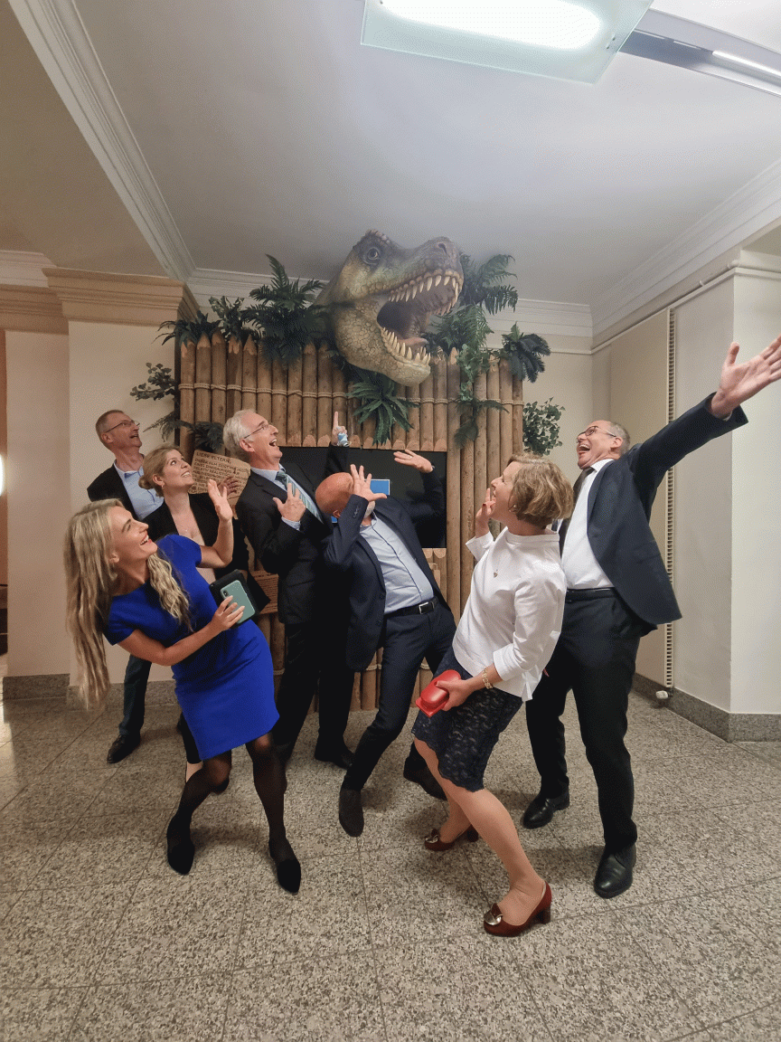 Gruppe von 7 Personen in Abendkleidung posiert erschrocken vor einem dekorativen T-Rex-Kopf an der Wand.