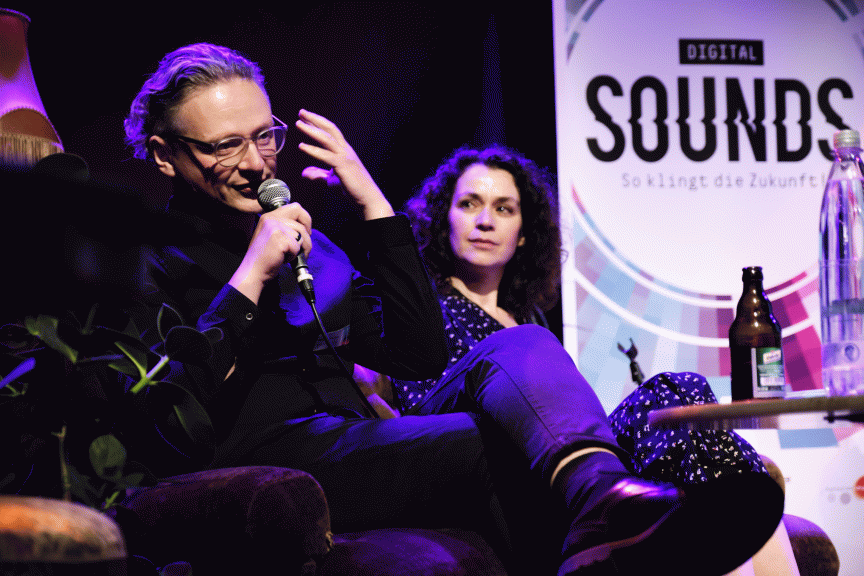Zwei Personen sitzen auf einer Bühne bei „Digital Sounds". Eine spricht ins Mikrofon, im Hintergrund ein Banner.