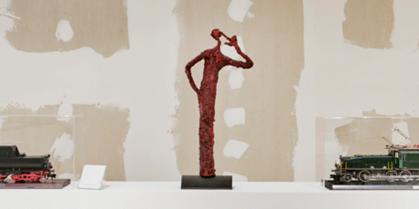 Rote abstrakte Metallskulptur einer menschlichen Figur auf schwarzem Sockel, flankiert von zwei Modelllokomotiven.