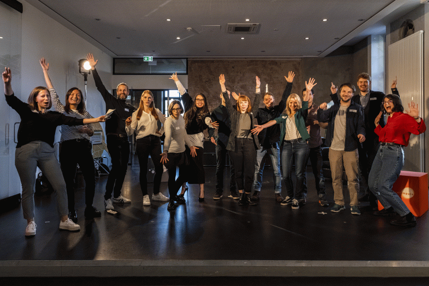 Gruppe von etwa 14 Personen steht auf einer Bühne, viele heben die Arme, lächeln in die Kamera.