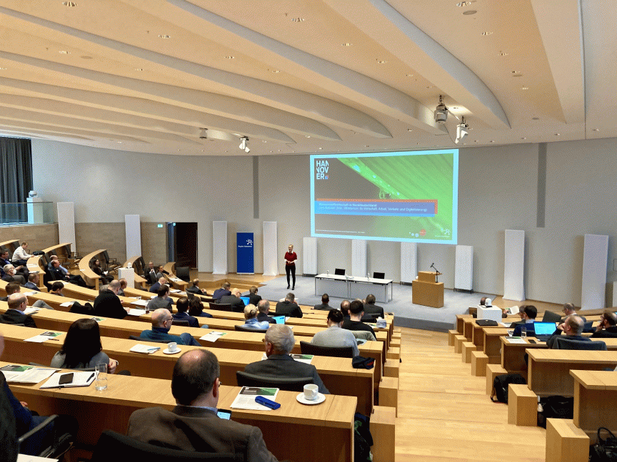 Vortragende Person steht vor einer Leinwand mit „Hannover"-Präsentation in einem modernen Hörsaal mit Publikum.