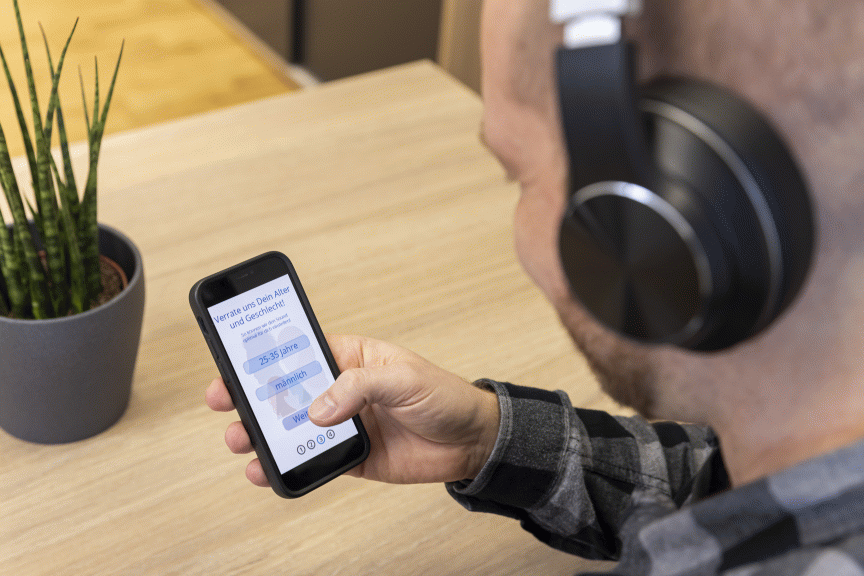 Person mit Kopfhörern hält Smartphone mit deutscher App-Oberfläche. Im Hintergrund Holztisch und Topfpflanze.